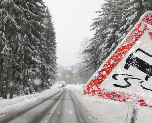 Auf einer verschneiten Straße befinden sich rechts und links verschneite Tannen und im Vordergrund ein Warnschild für Rutschgefahr